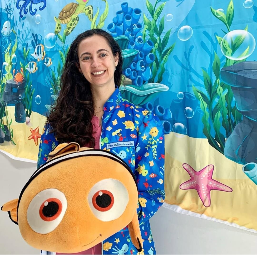 A Dra. Aline Bernardino sorrindo, de jaleco azul estampado de peixinhos, abraçada a um Nemo de pelúcia, com um painel do fundo do mar atrás